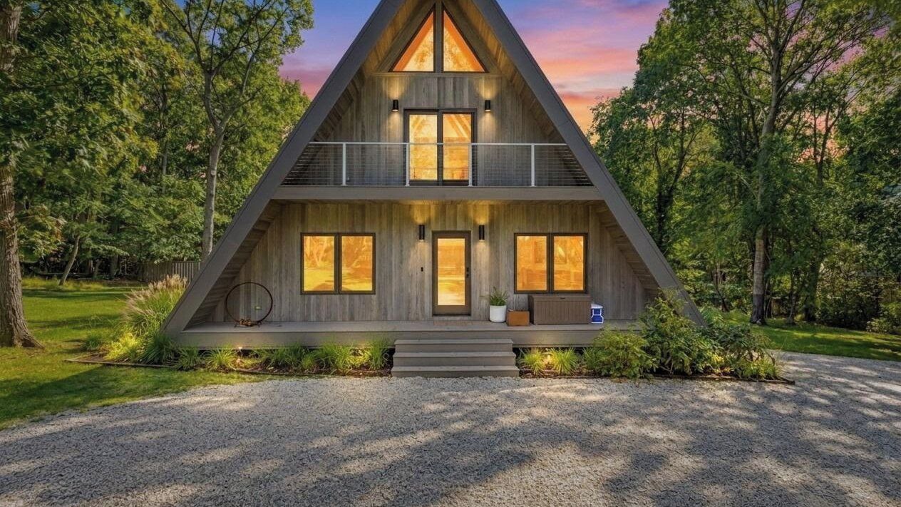 A modern, symmetrical, wooden cabin with a central entrance, large windows, and a balcony on the upper level.