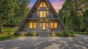 A modern, symmetrical, wooden cabin with a central entrance, large windows, and a balcony on the upper level.