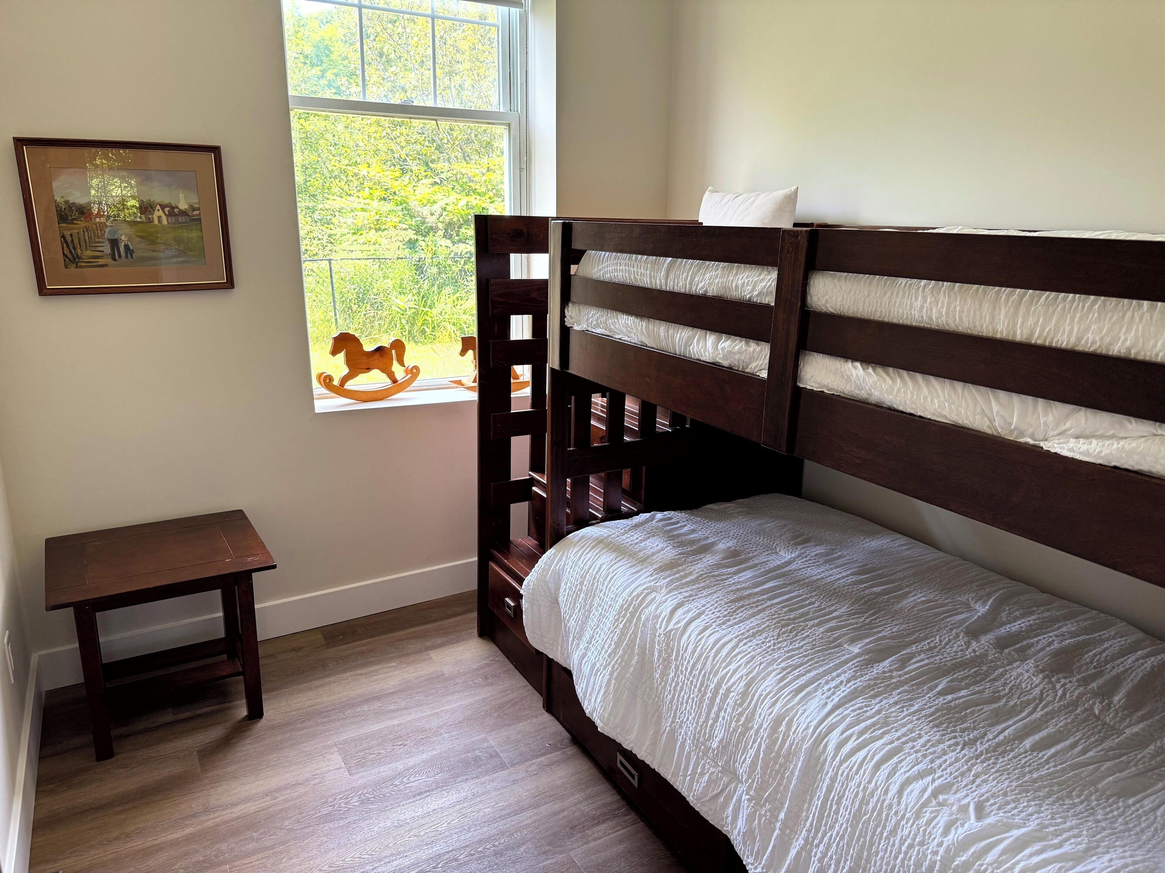 A small, neatly arranged bedroom with a wooden bunk bed, a small table, and a window with a view of greenery.
