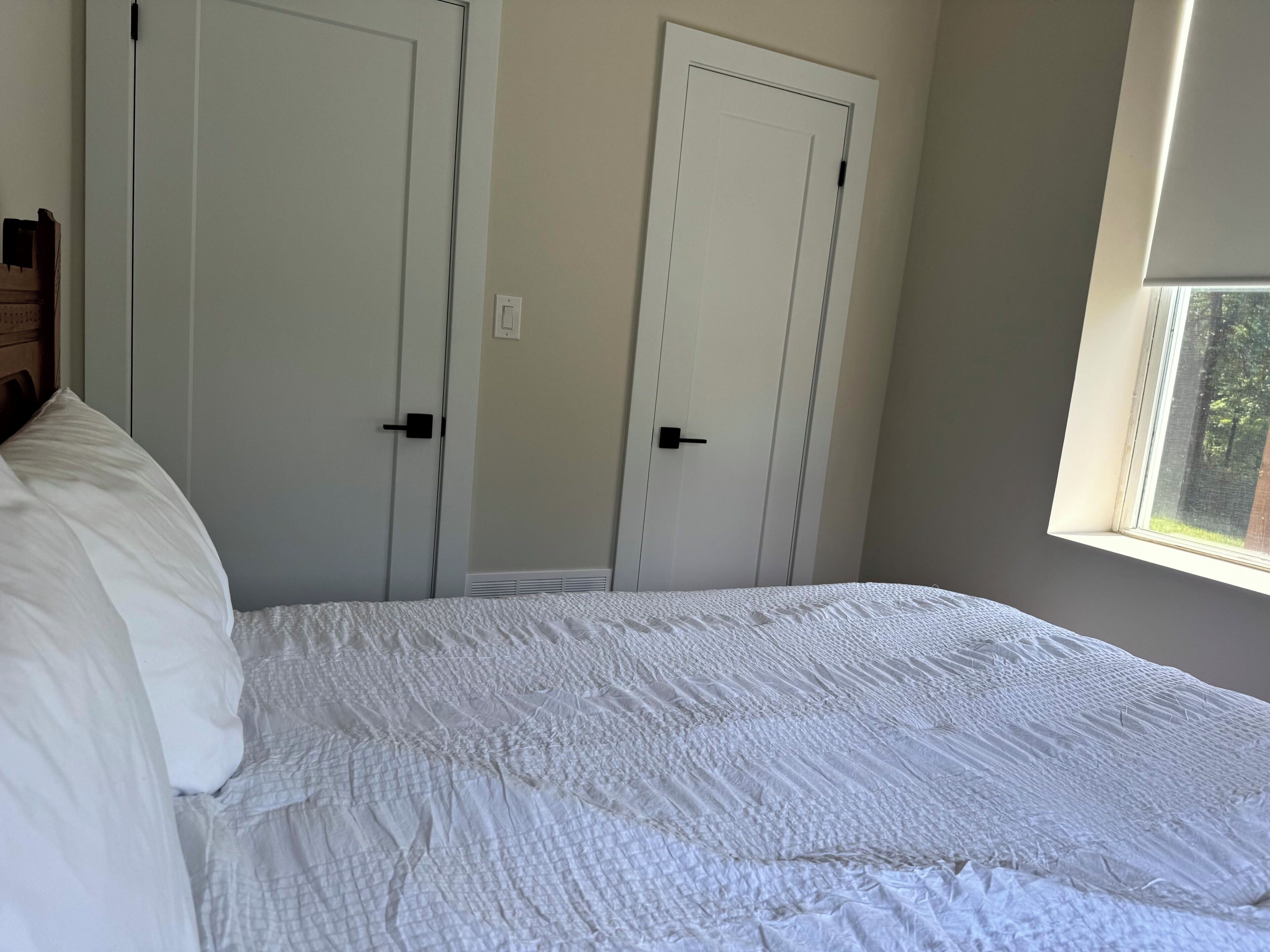 A bedroom with a bed, two white doors, and a window with a view of trees.