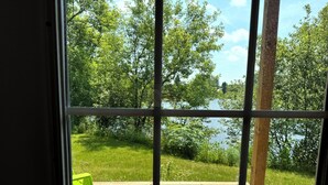 Blick durch ein Fenster auf eine Holzterrasse, grüne Stühle und einen von Bäumen umgebenen See.