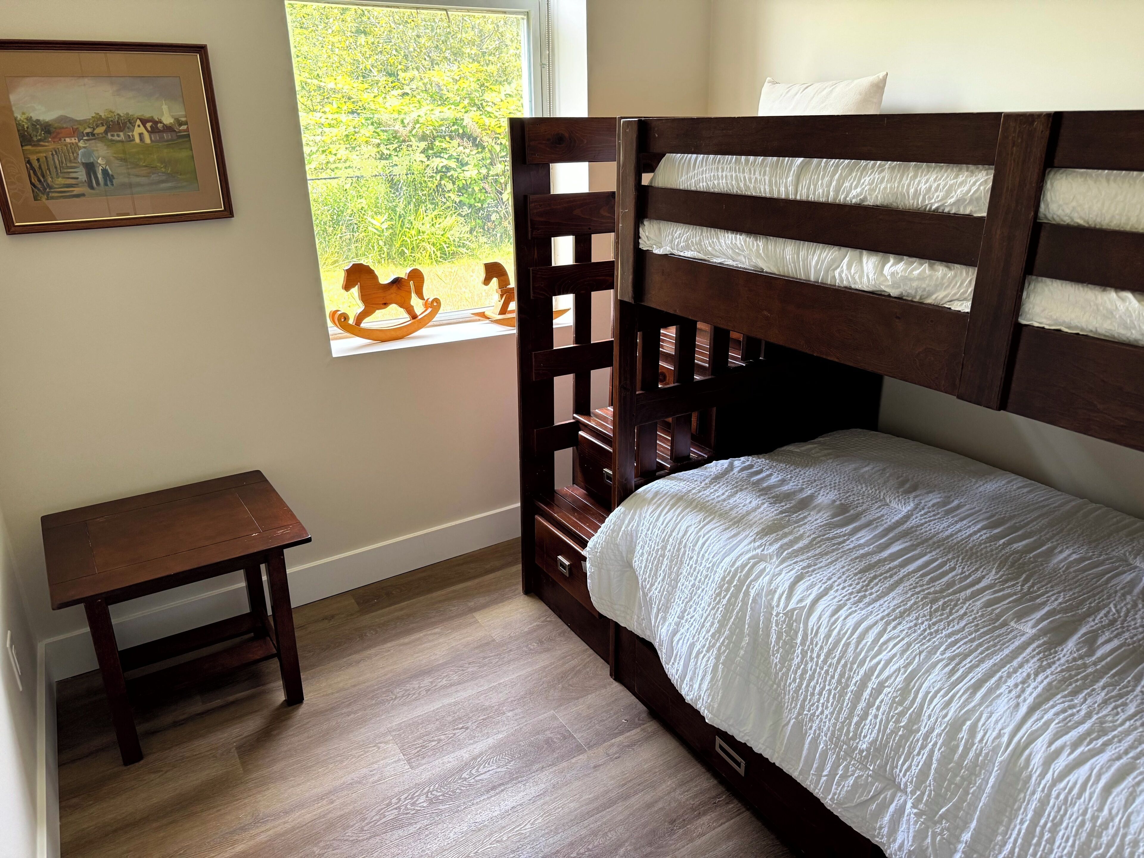 A bedroom with a bunk bed, a small wooden table, a framed painting, and a window with a view of greenery.