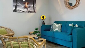 A living room with a blue sofa, a wicker chair, a table with a potted plant, a wall-mounted mirror, and a painting of a dog on a hammock.