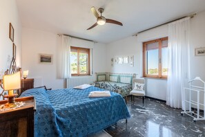 Une chambre à coucher comprenant un lit, une chaise, un bureau, un ventilateur de plafond et une fenêtre avec des rideaux.