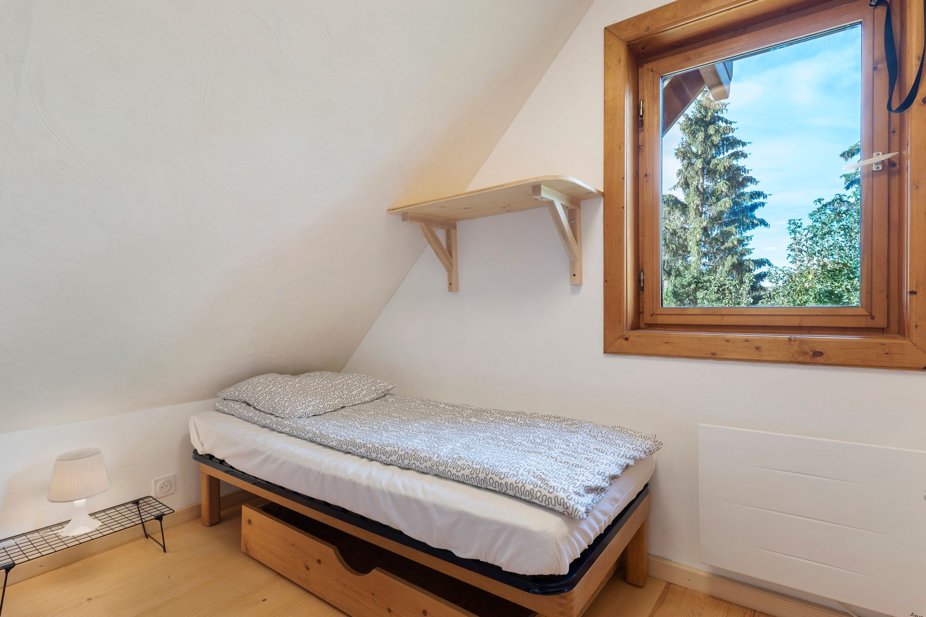 A small, single-bed room with a slanted ceiling, a window with a view of trees, and a wooden shelf above the bed.