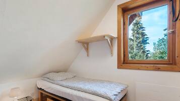 A small, single-bed room with a slanted ceiling, a window with a view of trees, and a wooden shelf above the bed.