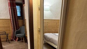A narrow hallway with wooden flooring, a bed with a white headboard, a chair, and a window with a curtain. - Apartment 'Maribel' with Shared Terrace and Wi-Fi (Pré-Saint-Didier)