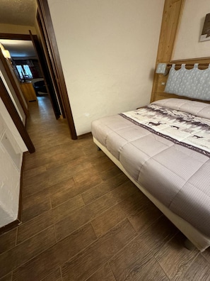 A bedroom with a wooden floor, a bed with a patterned bedspread, a wooden headboard, and a hallway leading to another room. - Apartment 'Maribel' with Shared Terrace and Wi-Fi (Pré-Saint-Didier)