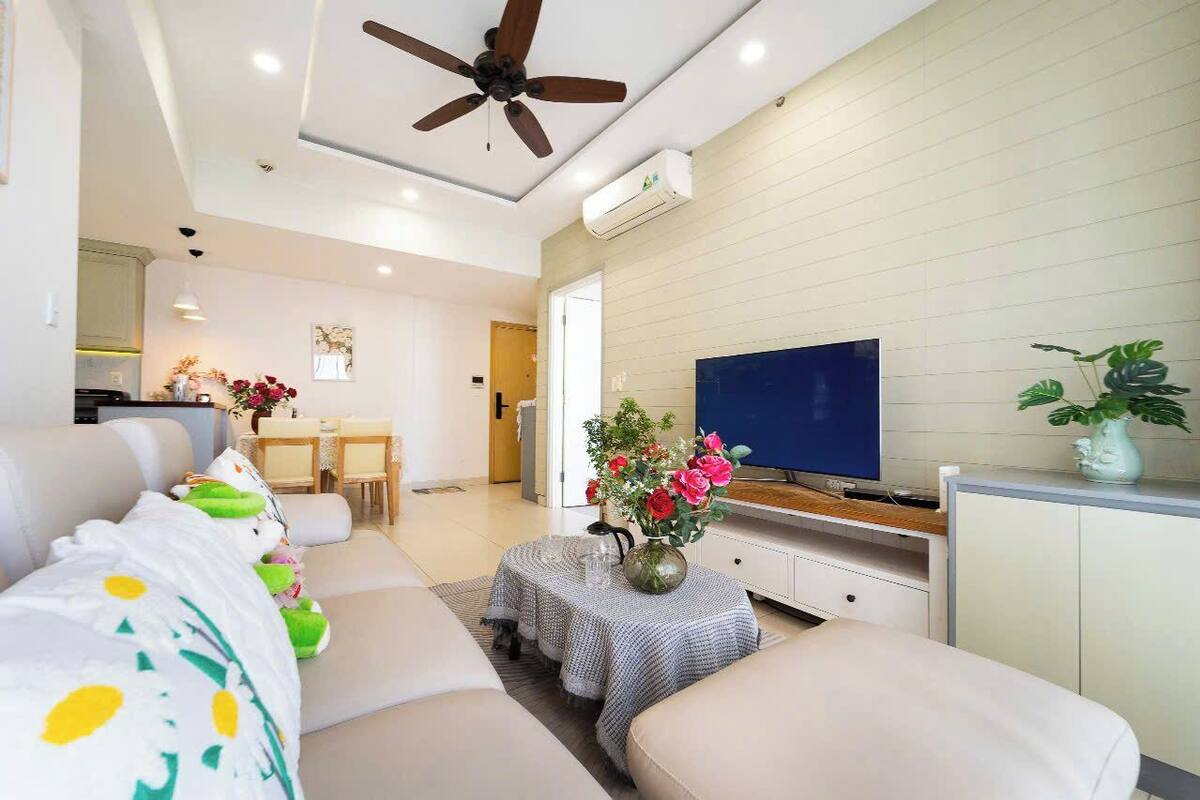 A modern living room with a sofa, a TV, a ceiling fan, and a dining area in the background.
