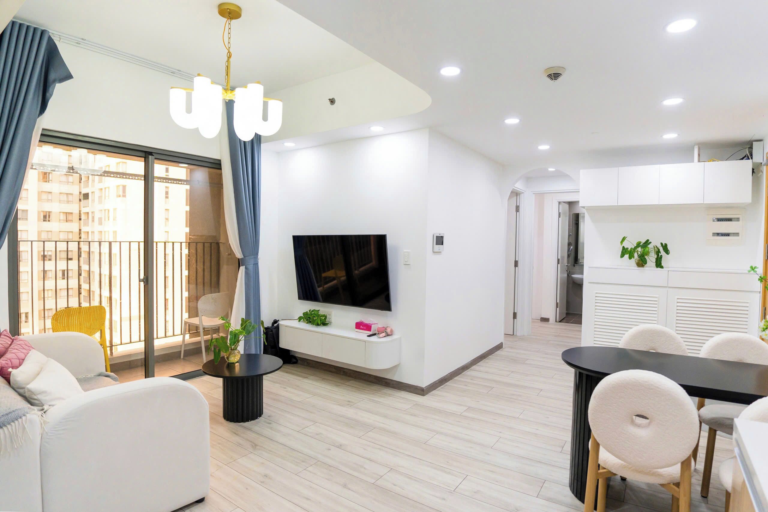 A modern living room with a flat-screen TV, a dining area with a black table and white chairs, and a balcony with a yellow chair.