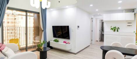 A modern living room with a flat-screen TV, a dining area with a black table and white chairs, and a balcony with a yellow chair.