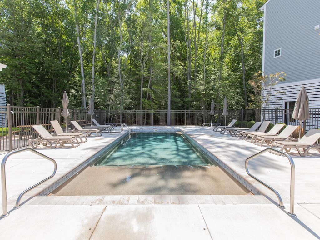 Ein rechteckiger Swimmingpool mit Metallgeländer, umgeben von Liegestühlen und Sonnenschirmen, vor einer Kulisse aus hohen Bäumen und einem Gebäude.