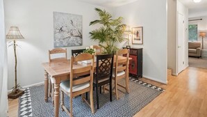 A dining area with a wooden table and chairs, a patterned rug, a potted plant, and artwork on the wall.