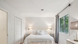 A bedroom with a bed, two bedside tables with lamps, a window with curtains, and a door.