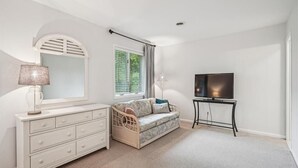 A bedroom with a white dresser, a wicker sofa, a television on a stand, and a window with blinds.
