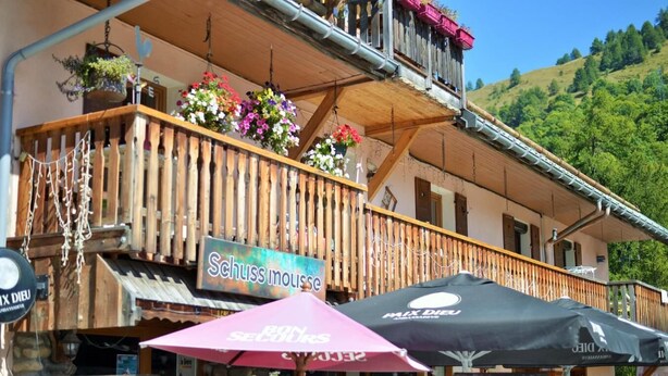 Un balcon en bois orné de jardinières suspendues, une enseigne indiquant « Schuss mousse », et des parasols d’extérieur.