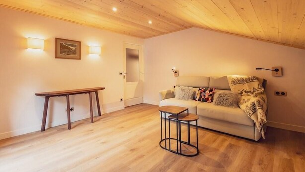 Un salón acogedor con techo de madera, un sofá con manta, una mesita y luces en la pared.