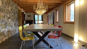 A dining area with a large wooden table, colorful chairs, a chandelier, stone walls, and large windows.