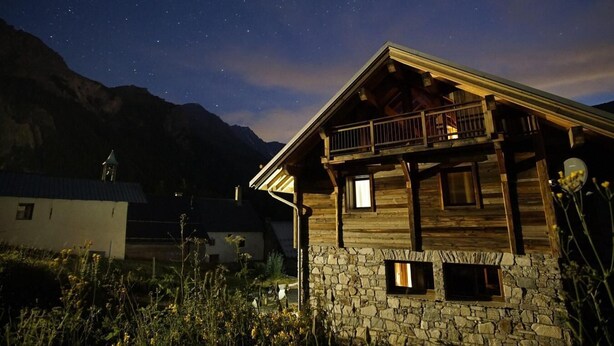 A wooden cabin with a stone foundation, a balcony, and a mountain backdrop.