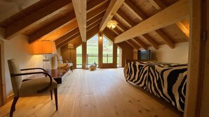 A spacious room with a high ceiling, wooden beams, a zebra-patterned bed, a desk with a lamp, and large windows offering a view of the outdoors.