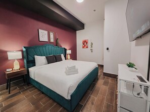 Ein Hotelzimmer mit einem wasserfarbenen ausgestatteten Bett, weißer Bettwäsche, einem Nachttisch aus Holz mit einer Lampe, einem kleinen Tisch ebenfalls mit einer Lampe, einem an der Wand montierten Fernseher und einer Topfpflanze auf einem weißen Tisch.