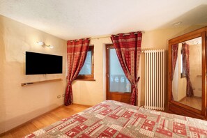 A bedroom with a bed, a TV mounted on the wall, a window with curtains, a radiator, and a wooden door.