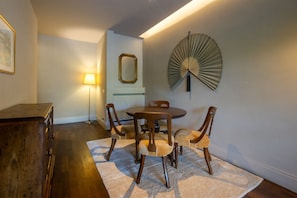 Un coin repas comprenant une table ronde et six chaises, un buffet en bois, une lampe sur pied et un ventilateur fixé au mur.