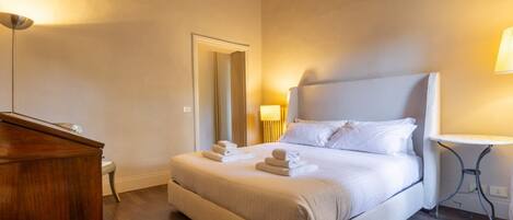 A neatly made bed with white linens and towels, a wooden dresser, a small table with a lamp, and a mirror on the wall.