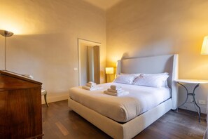 Un lit bien fait, avec du linge de lit et des serviettes blanches, une commode en bois, une petite table avec une lampe et un miroir accroché au mur.