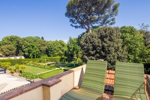 Una terraza con vista a un jardín cuidado, un árbol grande y un edificio con techo de tejas.