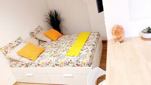 A neatly made bed with floral bedding, a yellow blanket, and several pillows. There is a small potted plant and a salt lamp on a wooden table beside the bed.