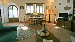 Una sala de estar con un sofá verde, una mesa de comedor con sillas, un televisor sobre un mueble de madera y un suelo de baldosas.