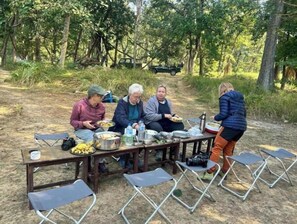 A group of people dining outdoors with a table set with food and drinks, surrounded by trees.