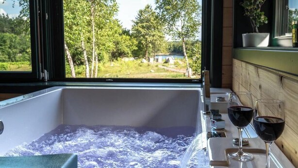 Eine Badewanne mit Schaum, zwei Gläser Rotwein und Blick auf einen See und Bäume durch ein Fenster.
