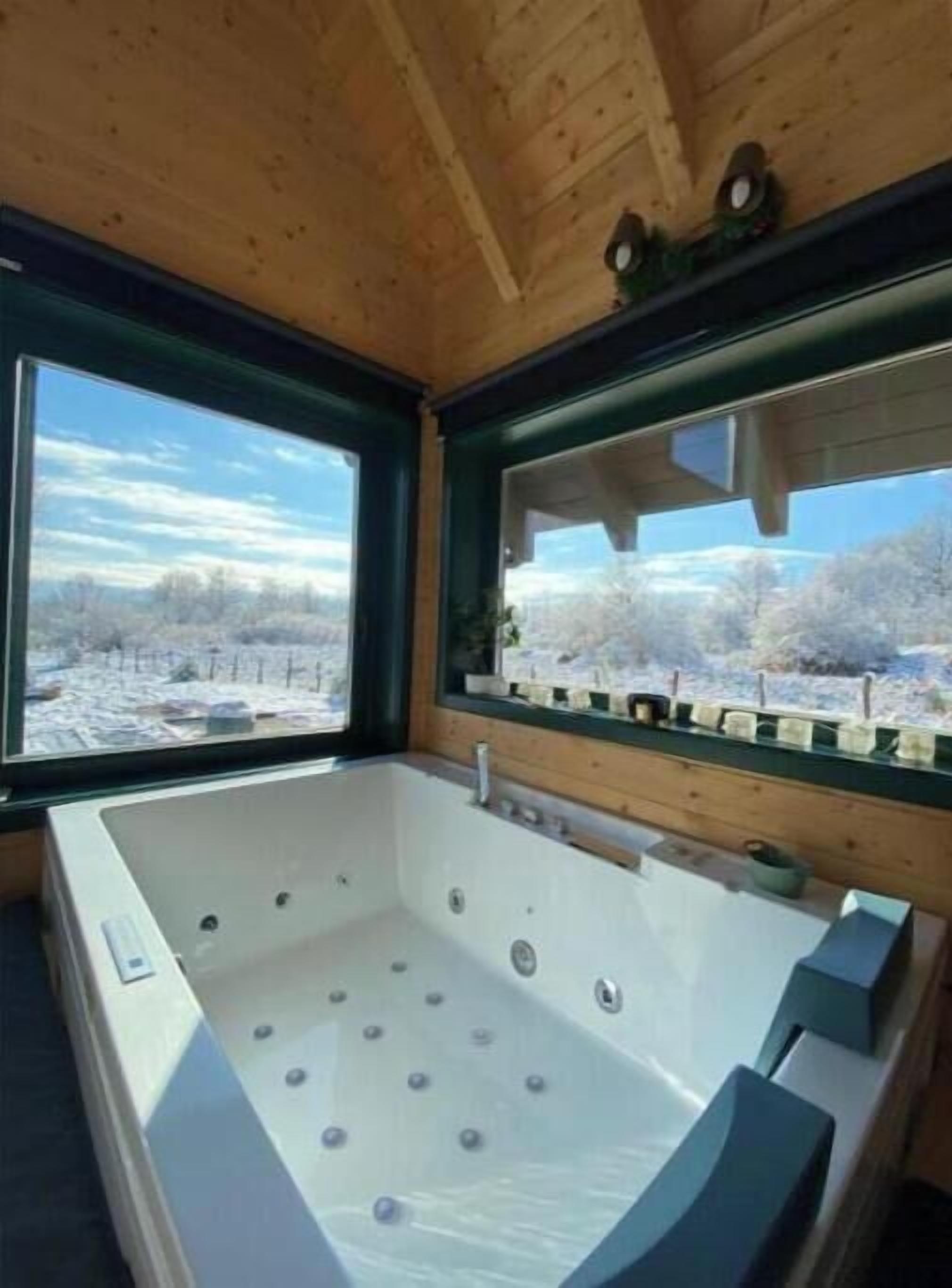 A wooden cabin interior with a large bathtub, wooden ceiling, and a window offering a view of a snowy landscape.