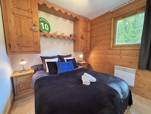 Quarto com painéis de madeira, cama, mesas de cabeceira, aquecedor e janela com vista para árvores.