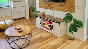 A modern living room with a flat-screen TV mounted on a wooden panel wall, a ceiling fan, a wooden cabinet with storage, a round coffee table, and a large plant. - Modern Home w/ Theater, BBQ & Private Yard (Ladson)