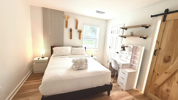 A bedroom with a bed, bedside tables, a vanity with a mirror and lights, a wooden wardrobe, and a window with blinds. - Modern Home w/ Theater, BBQ & Private Yard (Ladson)
