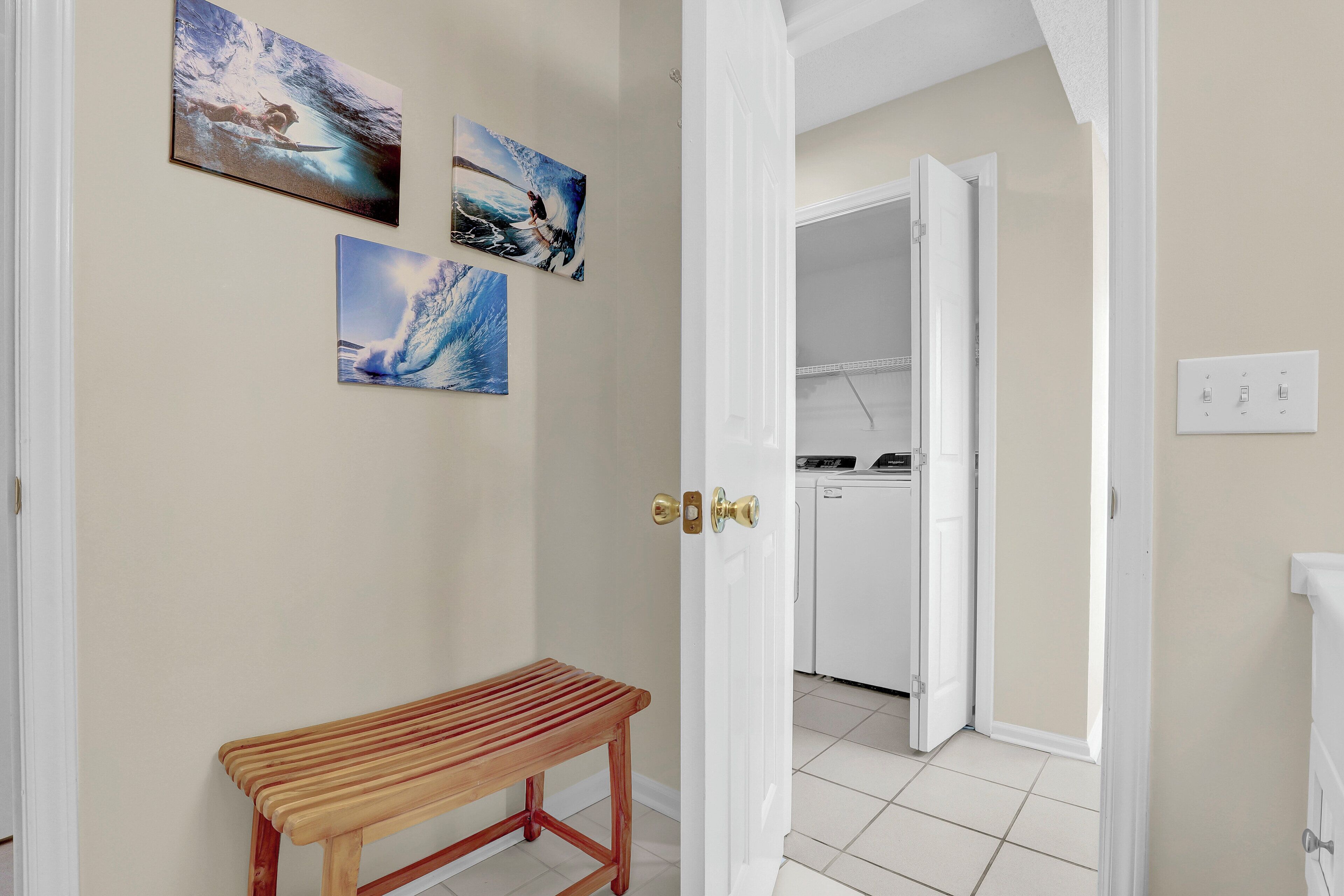 A hallway with a wooden bench, a door leading to a kitchen, and three wave-themed paintings on the wall.