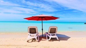 Deux chaises de plage sous un parasol rouge, sur une plage de sable fin, avec une eau turquoise et limpide et un voilier qui se profile à l’horizon.