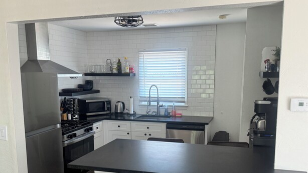 Una cocina compacta con paredes de azulejos blancos, un refrigerador de acero inoxidable, un microondas, una estufa y una cafetera. Hay dos sillas marrones y una ventana con persianas.