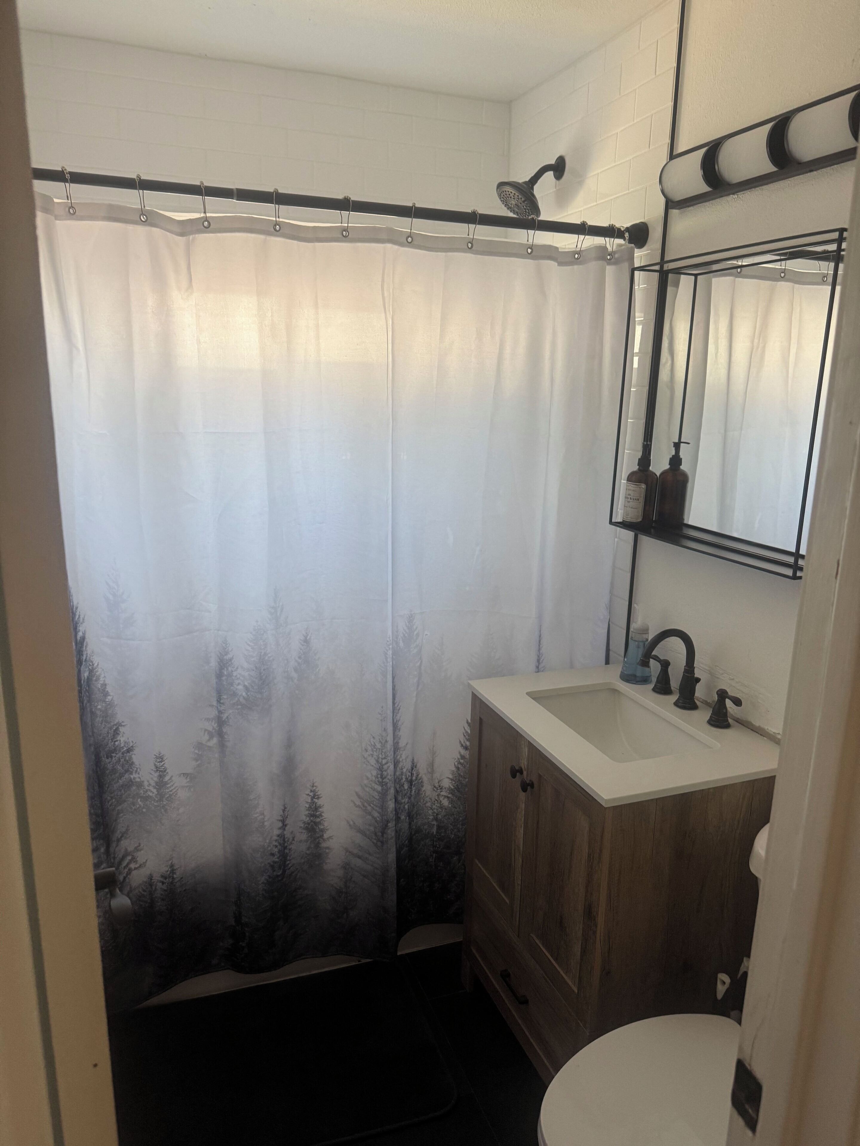 Baño con cortina de ducha mostrando un paisaje boscoso, tocador de madera con lavamanos y espejo sobre él.