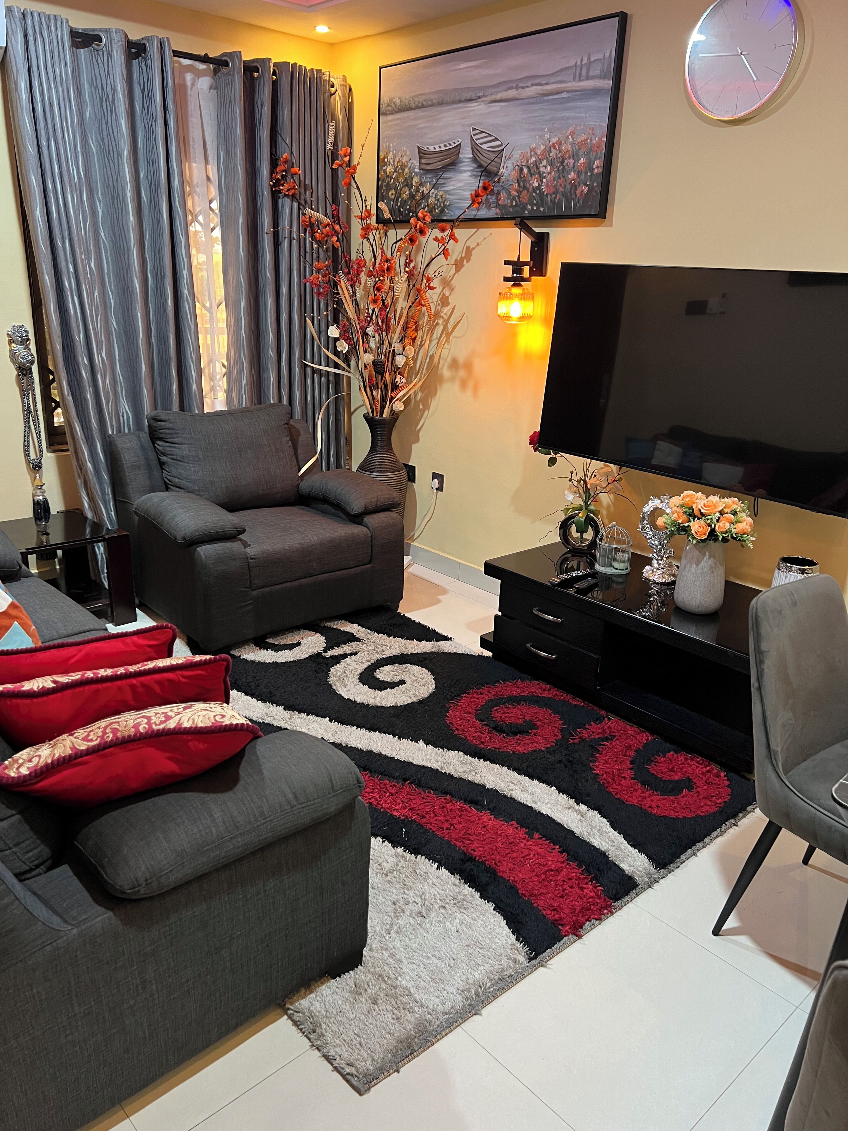 Ein Wohnzimmer mit grauem Sofa, einem roten und schwarzen Teppich mit Muster, einem Fernseher und einer Vase mit Blumen.