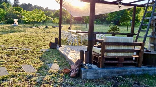 A wooden pergola with a cushioned bench and a table, set in a grassy outdoor area with a view of a forested landscape.