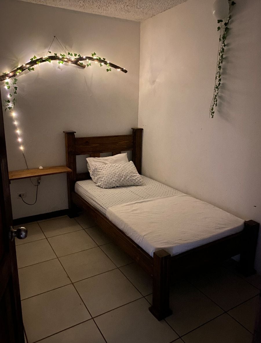 Una cama individual con cabecera de madera en una habitación con paredes blancas y suelo de baldosas.