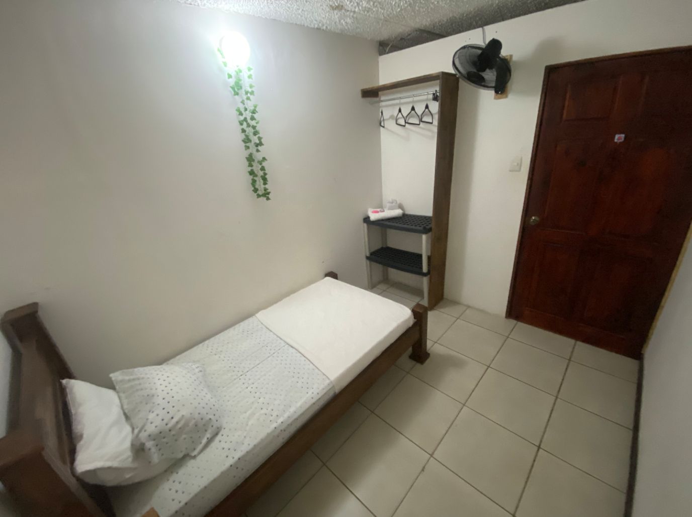 A small, single-bed room with a wooden bed frame, a white mattress, and a white pillow. There is a wooden door on the right and a wall-mounted shelf with a fan and a small item on it.