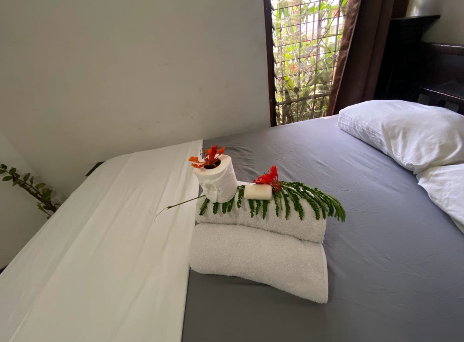 Un letto rifatto con un cuscino bianco, un asciugamano bianco arrotolato e un bouquet decorativo sul comodino.