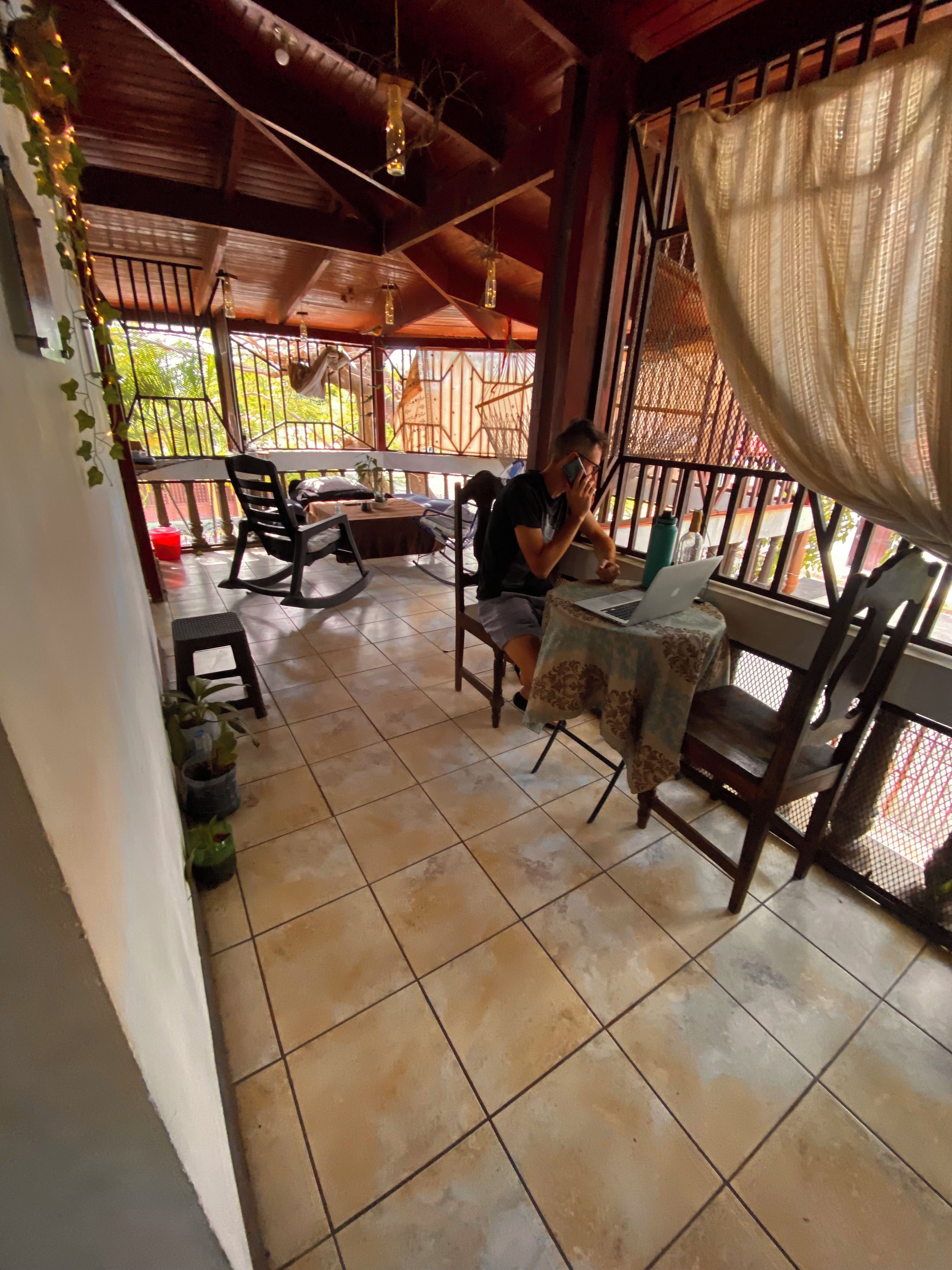A covered patio area with a table and chairs, a person using a laptop, and potted plants.