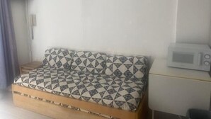 Ein kleines, kompaktes Zimmer mit einer Schlafsofa mit geometrischem Muster, einer Mikrowelle auf einem kleinen Tisch und einer schlichten weißen Wand.