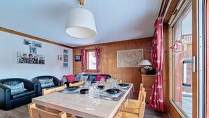Une pièce chaleureuse avec une table à manger en bois, préparée pour quatre personnes, un canapé en cuir noir et une grande fenêtre ornée de rideaux rouges.